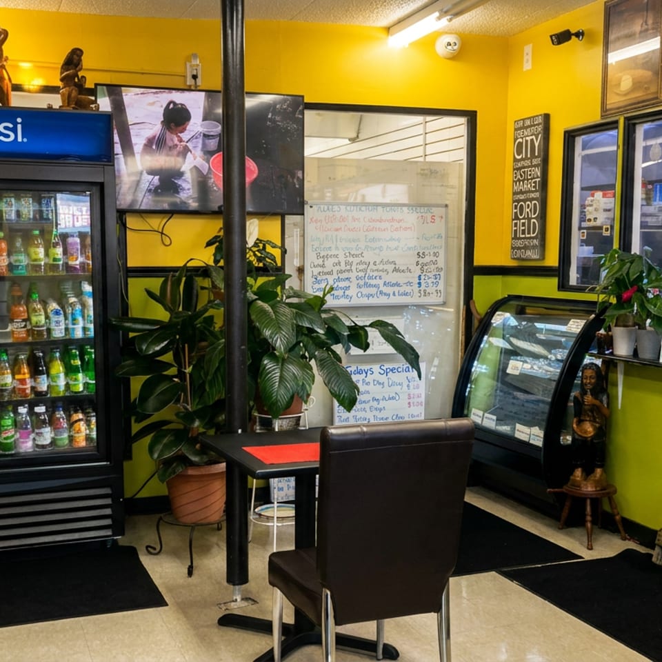 Colorful restaurant interior with yellow walls, beverage cooler, dining tables, plants, artwork, and a staff member behind the counter.